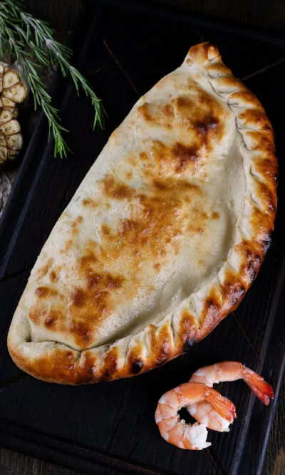 Запечене кальцоне з морепродуктами на дерев'яній дошці, прикрашене розмарином і креветками.